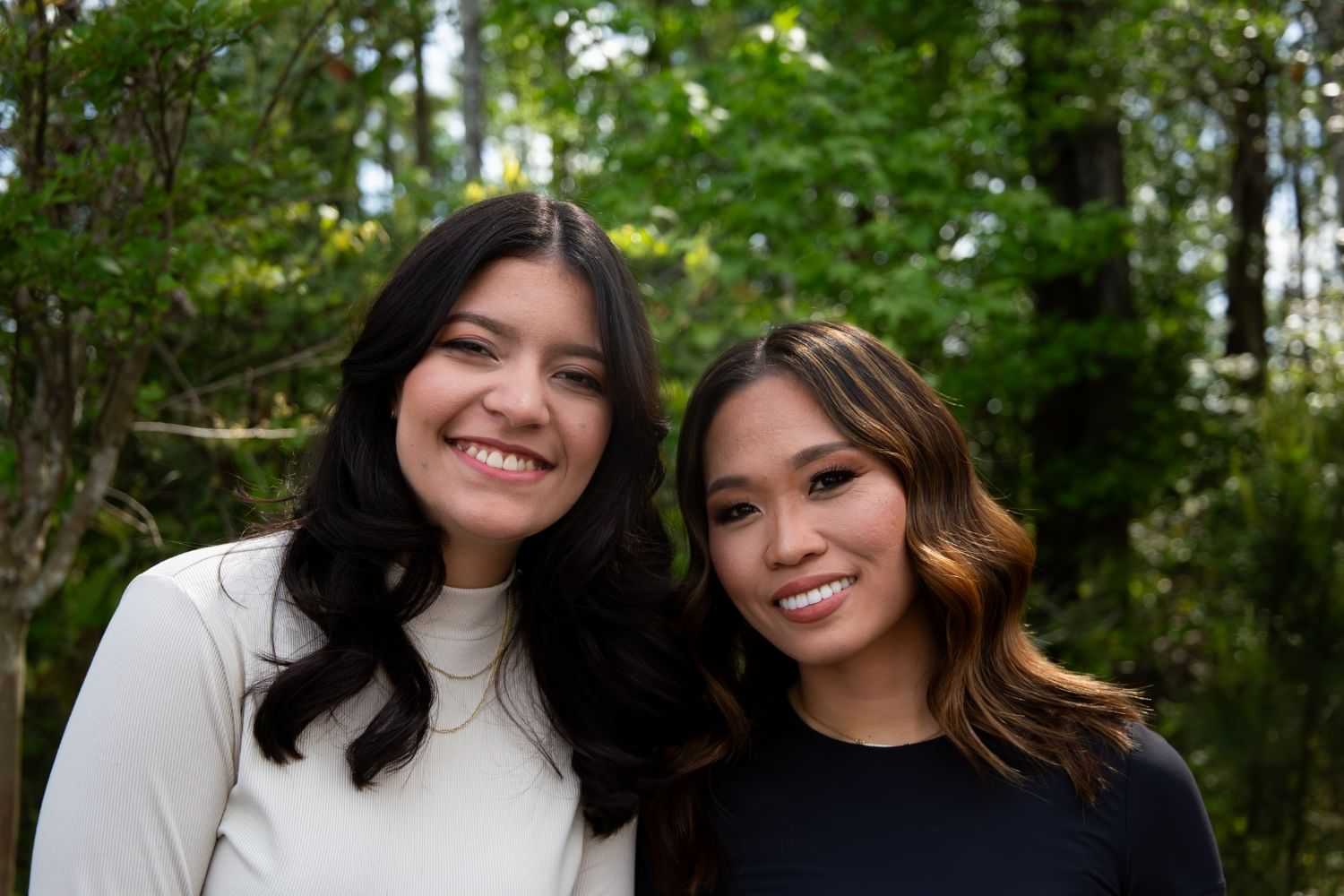 Two women smiling outdoors, surrounded by green trees and natural light.